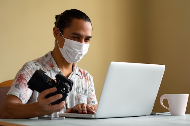 Il fotografo latinoamericano con maschera medica lavora a casa. Copia spazio. Concetto di coronavirus.