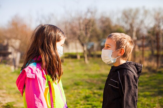 Il fratellino e la sorella stanno all'aperto in un giardino indossando maschere protettive che si guardano Protezione della famiglia durante l'epidemia Nuove regole di vita durante la quarantena