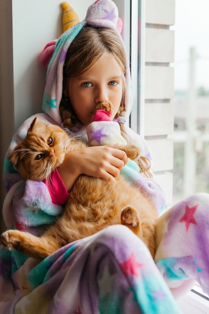 Il gattino dello zenzero e una bambina sono seduti sul davanzale della finestra e guardano fuori dalla finestra.