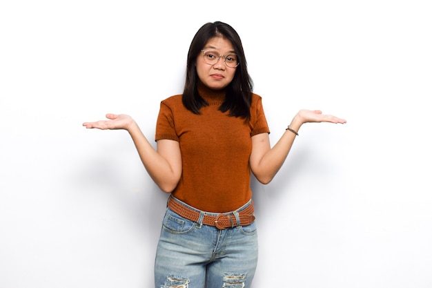Il gesto confuso di giovani belle donne asiatiche veste la camicia arancione isolata su fondo bianco