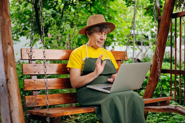 Il giardiniere anziano con un cappello sta guardando qualcosa su un computer portatile e sta effettuando una videochiamata nel cortile mentre è all'aperto