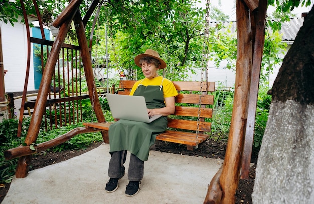 Il giardiniere anziano con un cappello sta guardando qualcosa su un computer portatile e sta effettuando una videochiamata nel cortile mentre è all'aperto