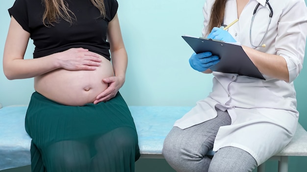 Il ginecologo della donna con la lavagna per appunti intervista la signora incinta con la pancia nuda prima della procedura che si siede vicino alla parete verde nel primo piano dell'ufficio della clinica