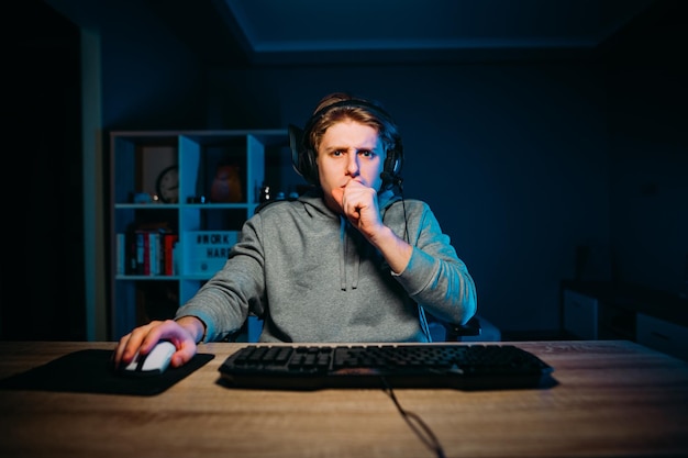 Il giocatore concentrato in una cuffia si siede a un tavolo a casa di notte e gioca ai videogiochi online