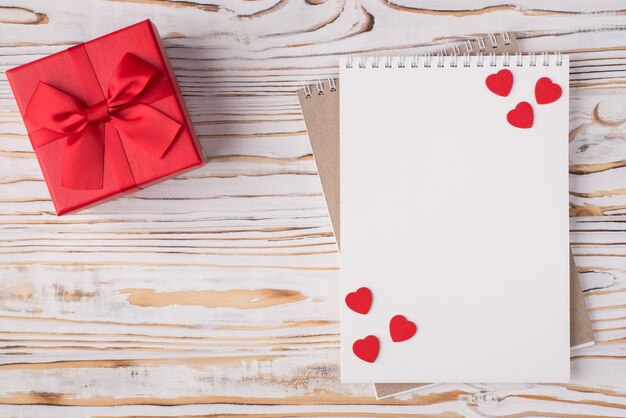 Il giorno di San Valentino concetto. In alto sopra la vista dall'alto foto piatta di un taccuino bianco presente rosso e cuori rossi isolati su sfondo di legno chiaro con copyspace