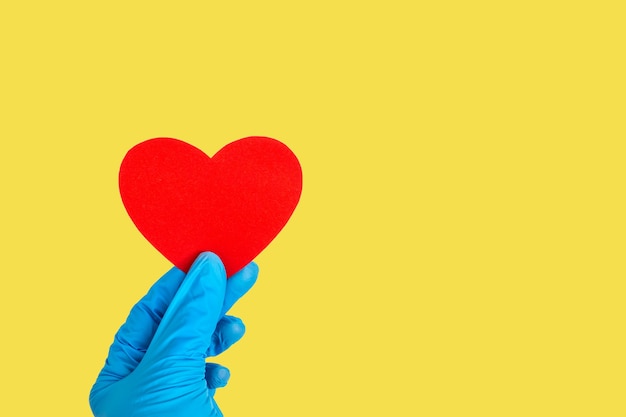 Il giorno di San Valentino concetto. Una mano in guanti blu medici tiene una forma di cuore rosso su uno sfondo giallo illuminante, copia spazio. Amore, concetto di salute