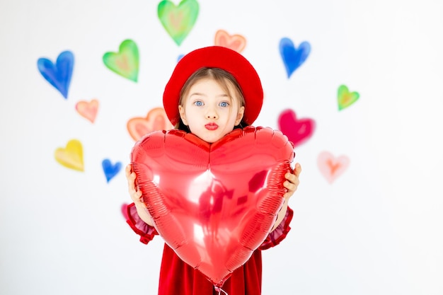 il giorno di San Valentino un piccolo bambino sorridente una ragazza con un berretto rosso felice tiene in mano un grande palloncino rosso a forma di cuore e si rallegra sullo sfondo di cuori colorati