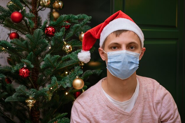 Il giovane a casa vicino a un albero di natale che indossa una maschera e un cappello di babbo natale guarda la telecamera