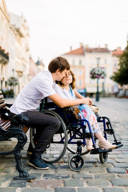 Il giovane abbraccia la sua donna sulla sedia a rotelle da dietro, mentre è seduto sulla panchina sulla strada della città vecchia