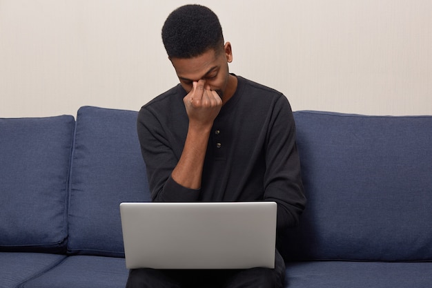 Il giovane adulto nero stressato infelice tiene la mano sul naso, cerca di concentrarsi, vestito con un maglione nero, lavora al computer portatile, si siede a un comodo divano, cerca di raccogliere informazioni