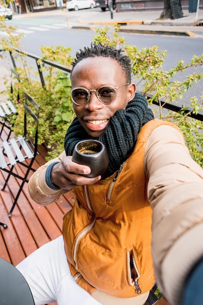 Il giovane africano si fa un selfie con il telefono bevendo caffè mentre è seduto fuori dalla caffetteria