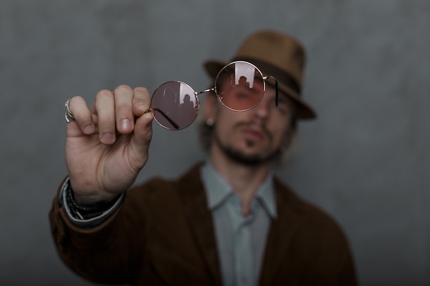 Il giovane alla moda con la barba in un cappello alla moda in abiti eleganti mostra alla fotocamera un paio di occhiali rosa rotondi vintage. Modello di ragazzo brutale nella stanza. Stile antiquato. Concentrati sugli occhiali.