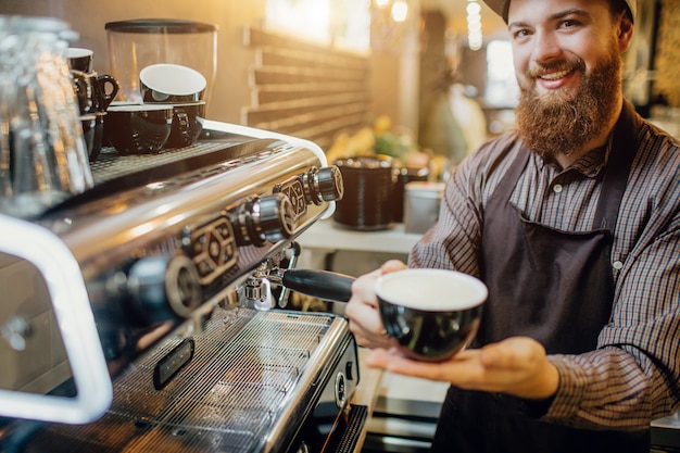 Il giovane allegro sta alla macchina del caffè e considera la macchina fotografica.