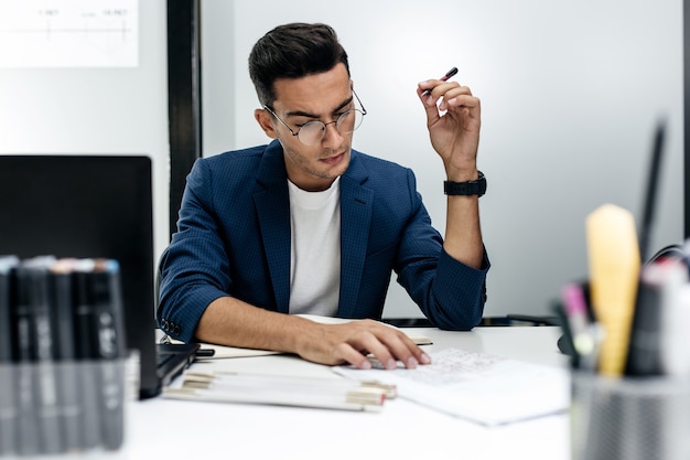 Il giovane architetto dai capelli scuri con gli occhiali e una giacca blu sta lavorando con i documenti sulla scrivania dell'ufficio.
