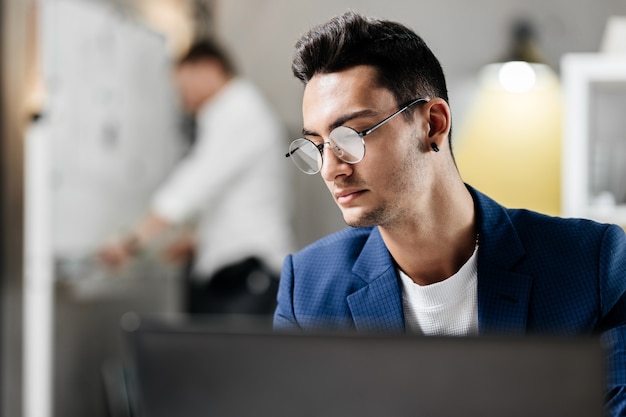 Il giovane architetto professionista con gli occhiali vestito con una giacca a scacchi blu lavora al computer portatile in ufficio.