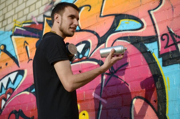 Il giovane artista dei graffiti con lo zaino e la maschera antigas sul suo collo dipinge i graffiti variopinti nei toni rosa sul muro di mattoni. Arte di strada e processo di pittura contemporanea