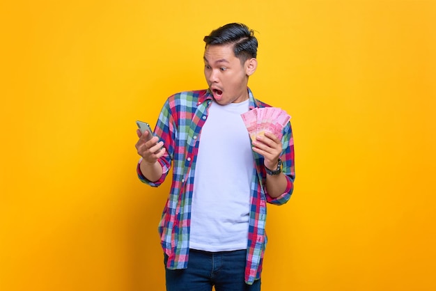Il giovane asiatico eccitato in camicia a quadri che tiene il telefono cellulare e le banconote in denaro ha ottenuto un prestito facile isolato su sfondo giallo
