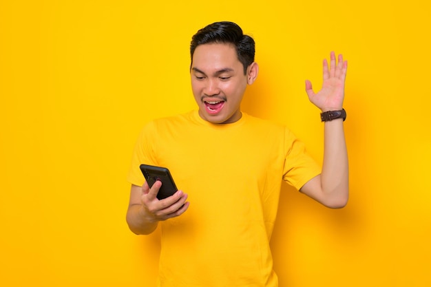 Il giovane asiatico eccitato in maglietta casual guardando il telefono cellulare si sente gioioso leggendo buone notizie isolate su sfondo giallo Concetto di stile di vita delle persone