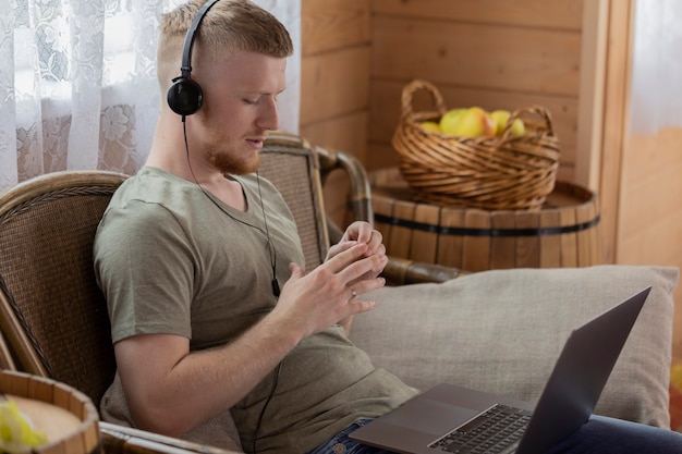 Il giovane attraente in cuffie lavora con il computer portatile, comunica nei social network, nella casa di campagna in legno