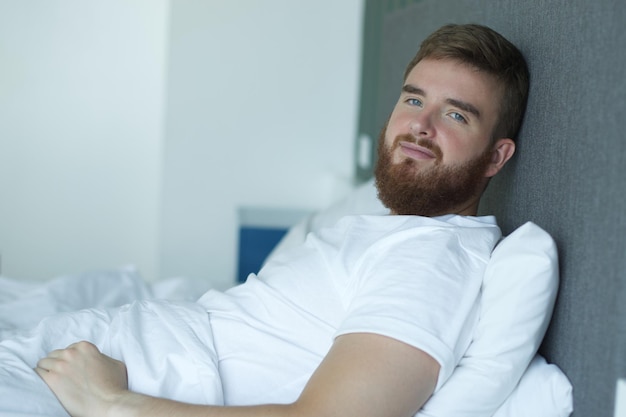 Il giovane bell'uomo assonnato con la barba felice giaceva a letto nella camera da letto a casa al mattino presto sorridendo e