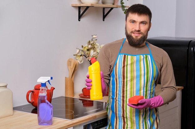 Il giovane bell'uomo barbuto in cucina, con indosso un grembiule e guanti rosa, pulisce la superficie di cottura usando detersivi
