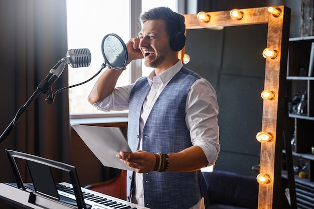 Il giovane bell'uomo barbuto sta ridendo in uno studio musicale di casa davanti a un pianoforte con uno specchio per il trucco dietro la schiena