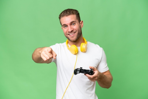 Il giovane bell'uomo che gioca con un controller per videogiochi isolato su sfondo verde chroma punta il dito contro di te con un'espressione sicura