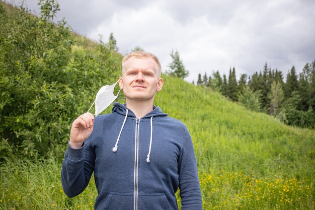 Il giovane biondo maschio europeo rimuove una maschera medica eliminabile in un parco su un fondo di una collina con erba e gli alberi.