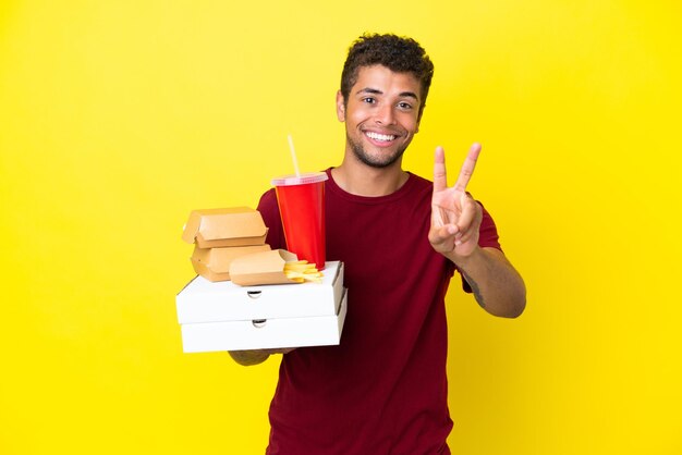 Il giovane brasiliano che tiene le pizze e gli hamburger ha isolato lo sfondo sorridente e mostra il segno della vittoria