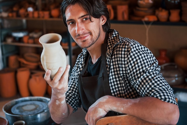 Il giovane ceramista tiene in mano un vaso fresco fatto a mano. Soddisfatto dei risultati del suo lavoro.