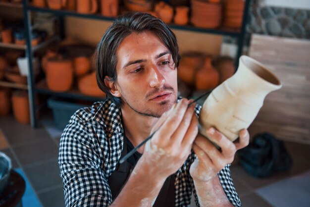 Il giovane ceramista tiene la sua pentola fresca fatta a mano e fa un po' di detallizzazione con uno speciale bastoncino.