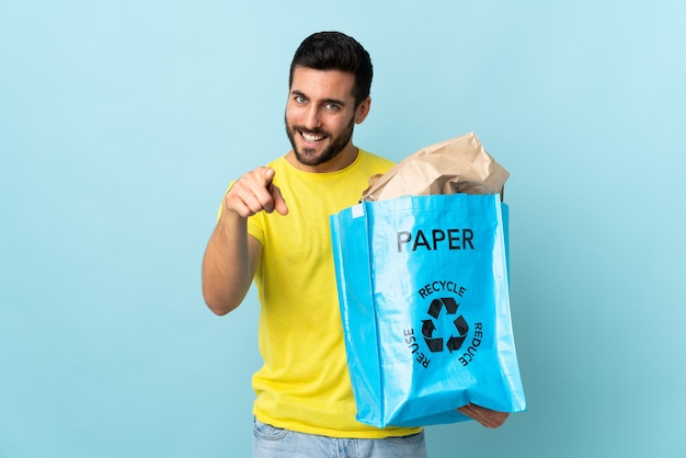 Il giovane che tiene un sacchetto di riciclaggio isolato sul muro blu punta il dito contro di te con un'espressione sicura