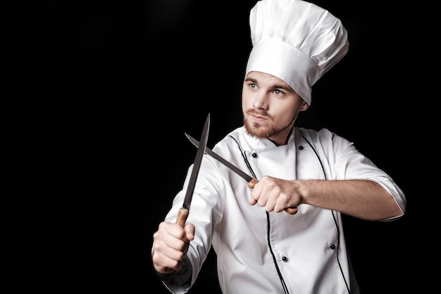 Il giovane chef barbuto in uniforme bianca tiene in mano due coltelli su sfondo nero