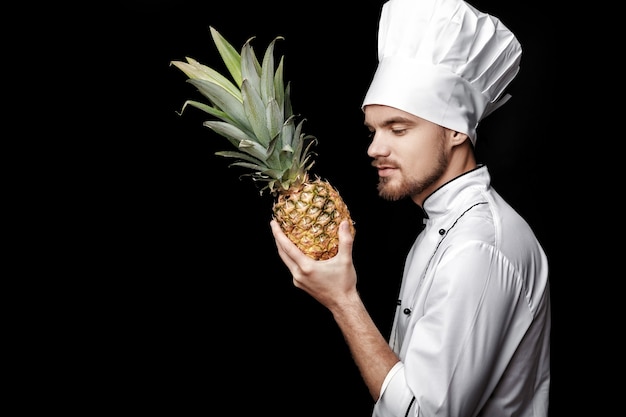 Il giovane chef barbuto in uniforme bianca tiene l'ananas fresco su sfondo nero