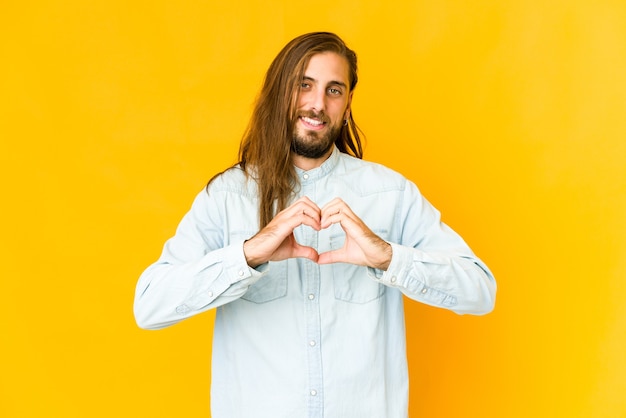 Il giovane con i capelli lunghi guarda sorridente e mostra una forma di cuore con le mani.