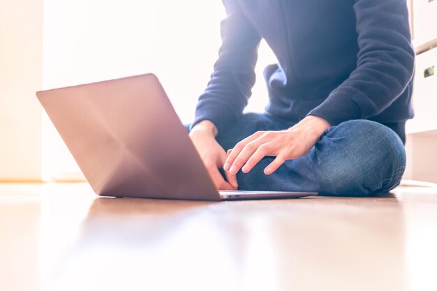 Il giovane con un computer portatile moderno sta sedendosi sul pavimento di legno