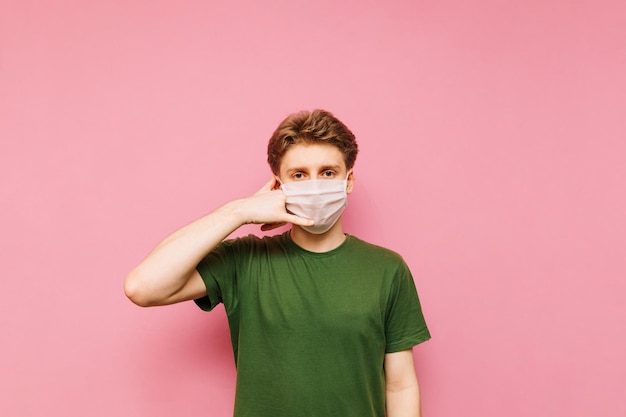 Il giovane con una maschera medica e abiti casual mostra un gesto di chiamata e guarda attentamente la fotocamera su uno sfondo rosa Pandemia di coronavirus COVID19 Quarantena