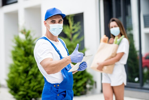 Il giovane corriere che indossa una maschera protettiva e guanti consegna merci a una giovane donna durante la quarantena