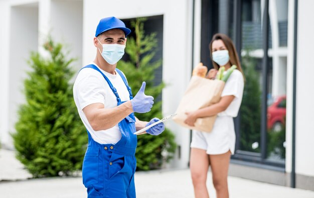 Il giovane corriere che indossa una maschera protettiva e guanti consegna merci a una giovane donna durante la quarantena