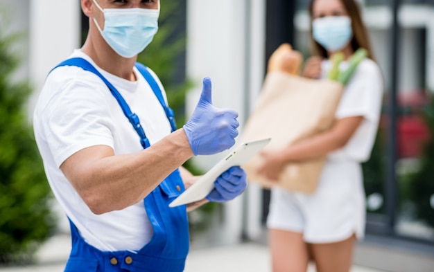 Il giovane corriere che indossa una maschera protettiva e guanti consegna merci a una giovane donna durante la quarantena