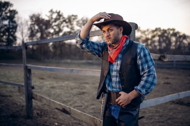 Il giovane cowboy in giacca di pelle e cappello pone contro il recinto di cavalli