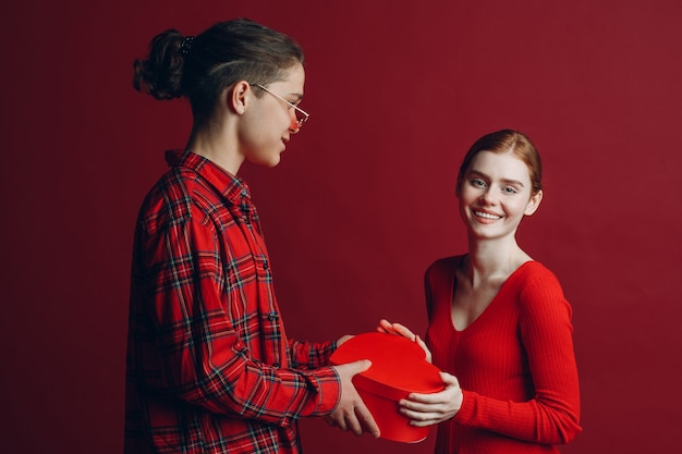 Il giovane dà alla sua ragazza una scatola a forma di cuore nel giorno di San Valentino su sfondo rosso studio