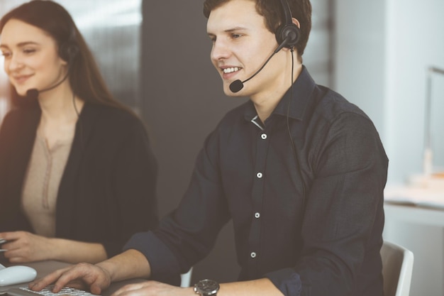 Il giovane dai capelli scuri con le cuffie sta parlando con un cliente, mentre è seduto in un ufficio soleggiato. Operatori di call center al lavoro