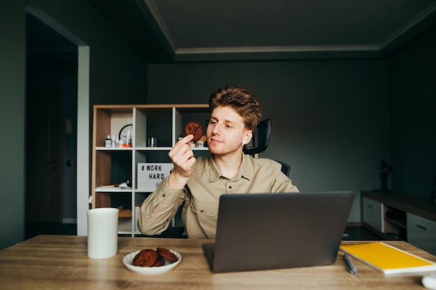 Il giovane divertente in camicia e cuffie wireless lavora a casa su un laptop sul posto di lavoro