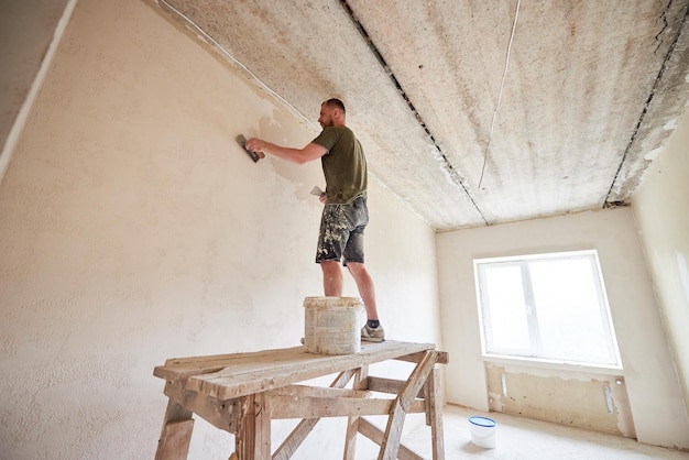 Il giovane è in piedi su un supporto di legno e lavora con una spatola con intonaco al muro contro una finestra