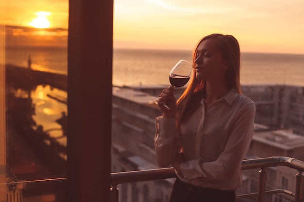 Il giovane è rilassante e bere un bicchiere di vino rosso sul balcone al tramonto la sera