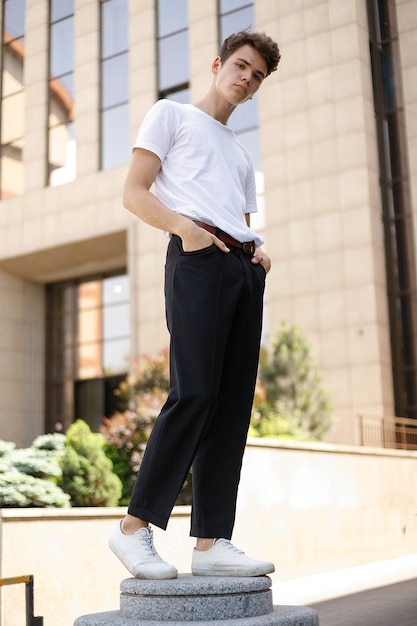 Il giovane elegante in una camicia nera alla moda in una t-shirt bianca alla moda in pantaloni neri con un'acconciatura alla moda riposa vicino a un moderno centro d'affari. Ragazzo attraente in strada in una giornata estiva.