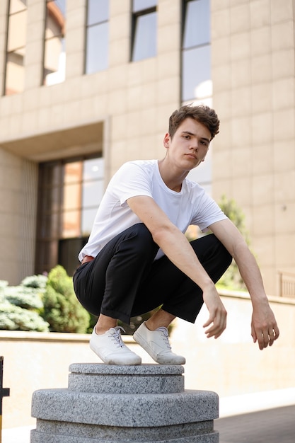 Il giovane elegante in una camicia nera alla moda in una t-shirt bianca alla moda in pantaloni neri con un'acconciatura alla moda riposa vicino a un moderno centro d'affari. Ragazzo attraente in strada in una giornata estiva.