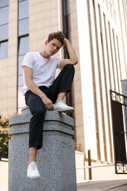 Il giovane elegante in una camicia nera alla moda in una t-shirt bianca alla moda in pantaloni neri con un'acconciatura alla moda riposa vicino a un moderno centro d'affari. Ragazzo attraente in strada in una giornata estiva.