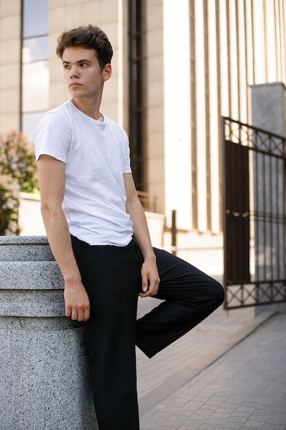 Il giovane elegante in una camicia nera alla moda in una t-shirt bianca alla moda in pantaloni neri con un'acconciatura alla moda riposa vicino a un moderno centro d'affari. Ragazzo attraente in strada in una giornata estiva.